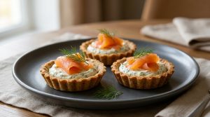 Trois tartelettes au blé noir garnies de crème aux herbes et de saumon fumé, servies sur une assiette grise.