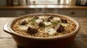 Plat à four en céramique contenant un crumble d'agneau et de chèvre fumant, garni de brins d'herbes aromatiques.
