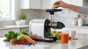 Main insérant une tranche de pomme dans un extracteur de jus Fridja F2500 entouré de fruits et légumes frais.