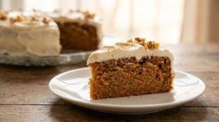 Une part de gâteau aux carottes nappée de glaçage blanc et décorée de noix sur une assiette blanche.