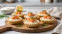 Cinq petites tartelettes garnies de fromage frais, saumon fumé et aneth sur une planche en bois ronde.
