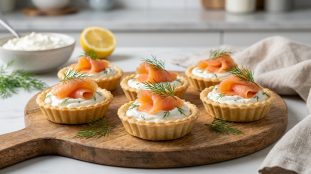 Cinq petites tartelettes garnies de fromage frais, saumon fumé et aneth sur une planche en bois ronde.